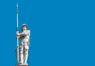 A stone statue of a medieval gatekeeper holding a spear upright. The clear sky is in the background. A stone statue of a medieval gatekeeper holding a spear upright. The clear sky is in the background.